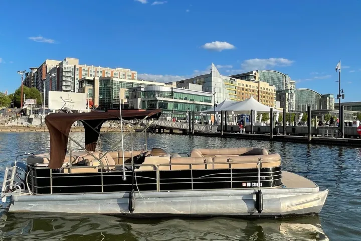 A pontoon boat on a city river with modern buildings and a clear sky in the background.